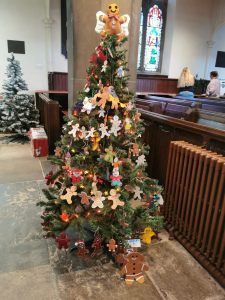 A Christmas tree standing in a traditional church. The tree is decorated with gingerbread man shapes, of different sizes, colours and materials. Another Christmas tree, and the church's stained glass windows are visible in the background.