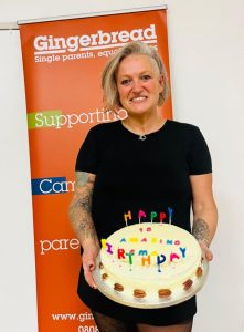 Ema is smiling and holding a cake with white icing and colourful happy birthday candles. She is standing in front of a Gingerbread pull up banner which reads 'Gingerbread: single parents, equal families'